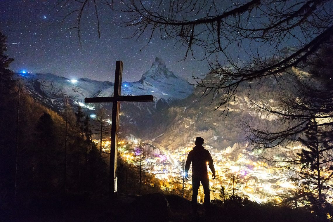 A man standing next to a cross with the backdrop of a mountain town lit at night