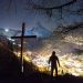 A man standing next to a cross with the backdrop of a mountain town lit at night